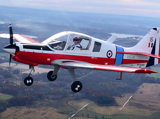 Scottish Aviation Bulldog aircraft in flight over rural landscape with pilot visible in cockpit