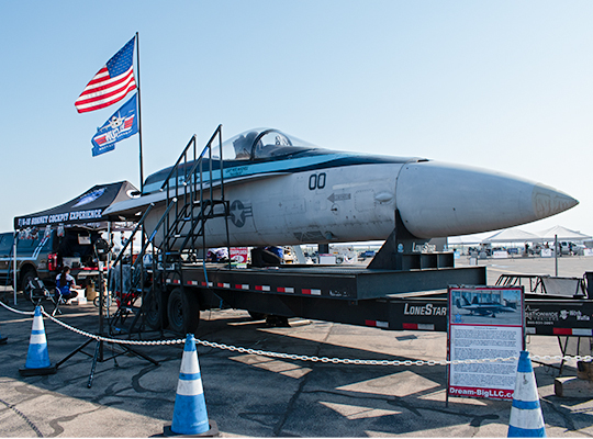 F-18 cockpit simulator mounted on trailer with stairs, ropes, and signage for interactive experience