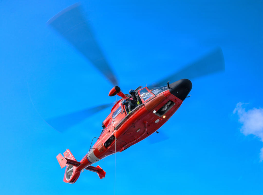 U.S. Coast Guard MH-65 Dolphin helicopter in bright red livery flying overhead against a clear blue sky, rotor blades blurred in motion, with crew visible in the cockpit.
