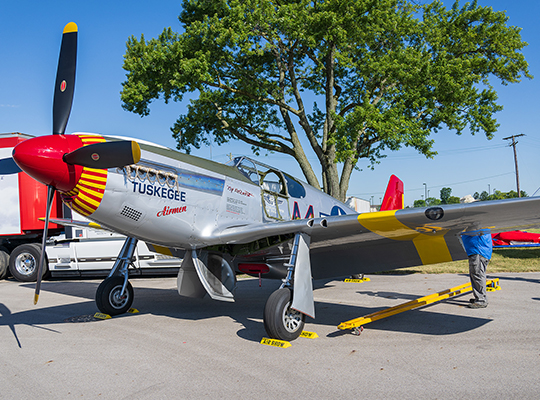 P-51 Mustang “By Request” parked on tarmac with red nose and Tuskegee Airmen markings, trees in background