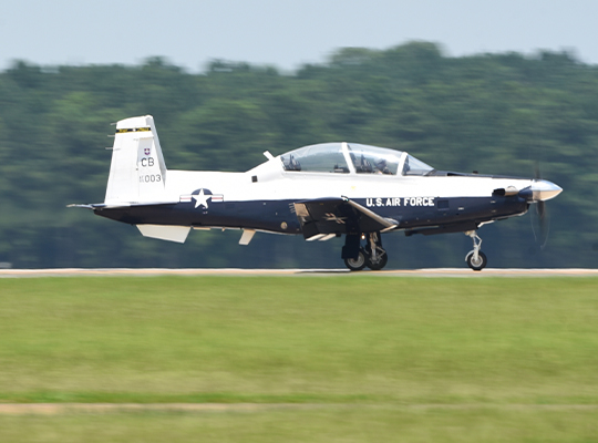 USAF T-6 Texan II training aircraft landing on runway with trees in the background