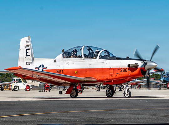 U.S. Navy T-6 Texan II training aircraft on display with orange and white paint scheme and spectators in background