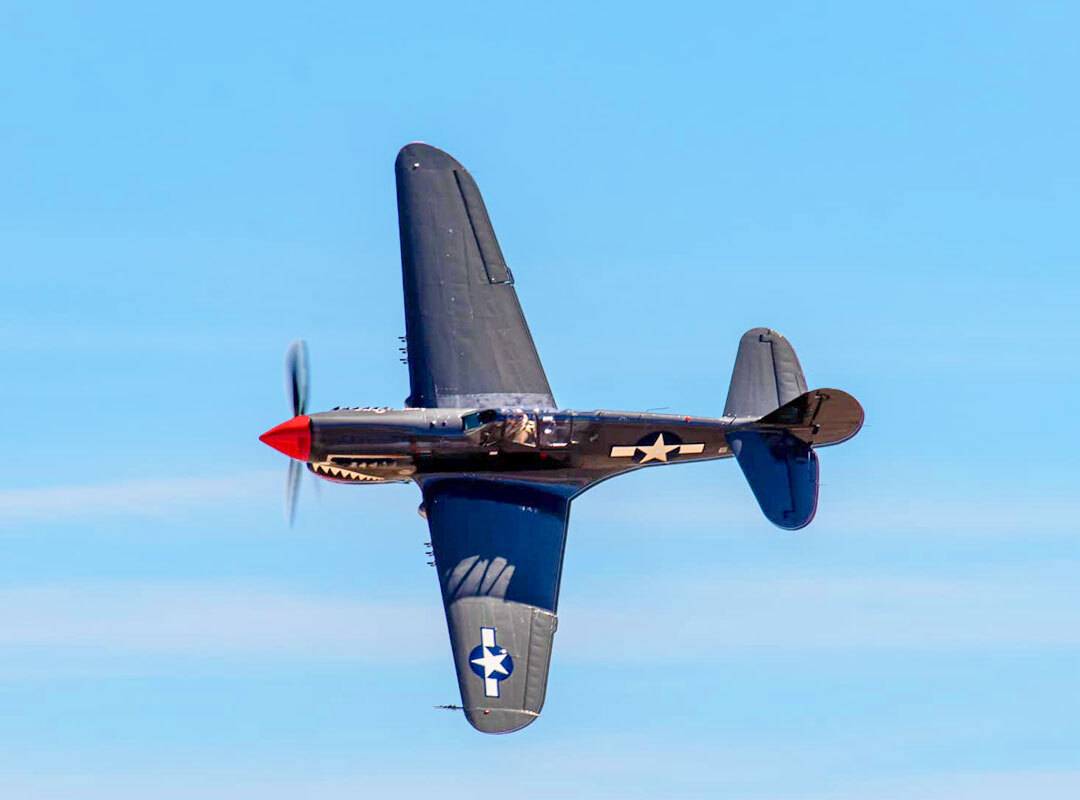 P-40 Warhawk fighter aircraft in flight, banking against a clear blue sky, featuring a red nose cone, shark mouth nose art, and U.S. military star insignia on the fuselage and wing.